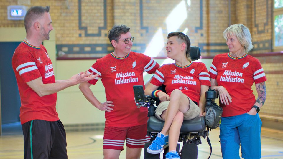 Mitglieder des Vereins Pfeffersport präsentieren in ihrer Sporthalle ihr neues Trikot mit der Aufschrift Mission Inklusion. Mit dabei ein Sportler, der im Rollstuhl sitzt.