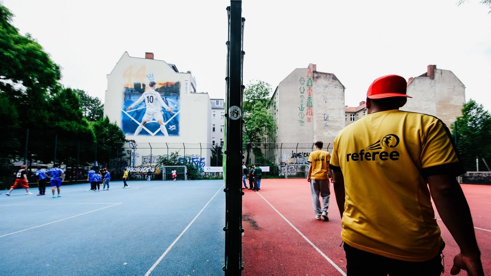 Das Foto zeigt eine urbane Fußball-Freifäche in Berlin. Im Hintergrund Wohnhäuser mit Fußballmotiv.